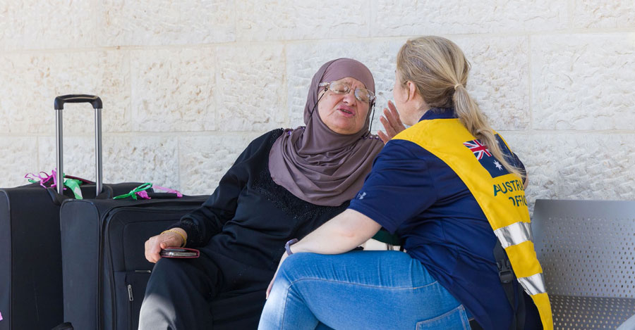 A seated conversation between an elderly female evacuee wearing a hijab and a female DFAT officer in a yellow Australian Official vest. The evacuees luggage nearby.