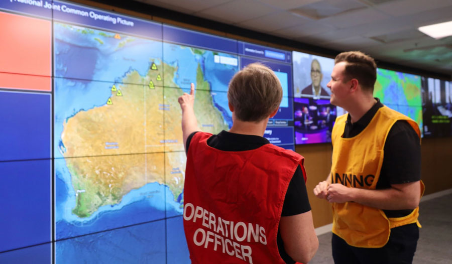 NEMA staff, dressed in operations officer vests, stand next to and point at a wall of screens. These screens display a map of Australia on the National Joint Common Operating Picture (NJCOP).