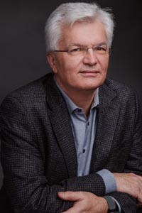 Middle aged man with white hair and wearing glasses faces camera. He wears a coat and a collared shirt.