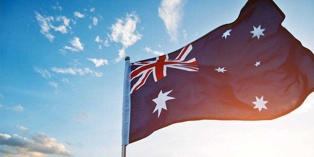 In the foreground is a mainly blue flag with white star shapes at right and left bottom. At top right is a red, white and blue design of crosses. The flag is attached to a white pole and in the background is a mainly blue sky.