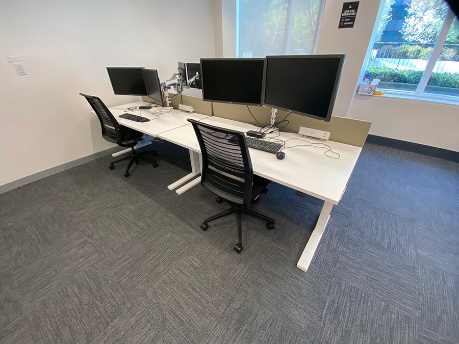 Shows a pod of desks in front of a window equipped with ergonomic chairs, double adjustable monitors, height adjustable desks and computer keyboards and mice.