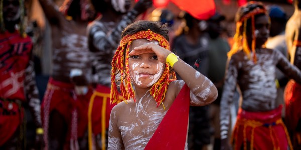 Young First Nations boy painted with ochre on his face and body, wearing a traditional red and yellow headpiece and a red ceremonial lap lap tied around his waist. 