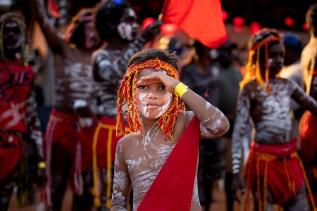 Young First Nations boy painted with ochre on his face and body, wearing a traditional red and yellow headpiece and a red ceremonial lap lap tied around his waist. 