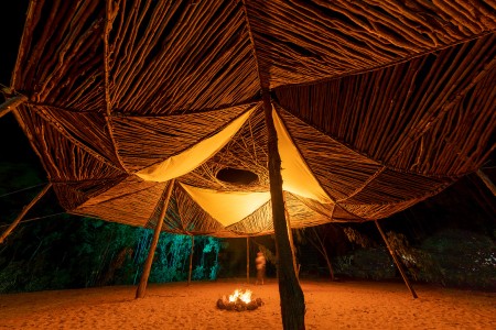 Night-time shot of Dhäwumi Wäŋa, the house of stories, with a string-bark eucalyptus structure and a fire in the centre.