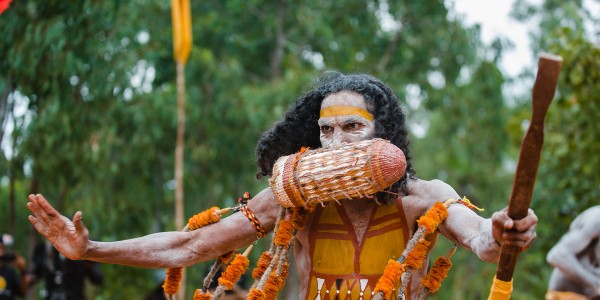 First Nations man with ochre paint on his face and body, performing or dancing with ceremonial accessories.