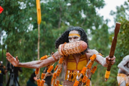 First Nations man with ochre paint on his face and body, performing or dancing with ceremonial accessories.