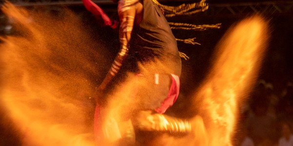 First Nations man with ochre body paint kicking up dust as he dances barefoot.