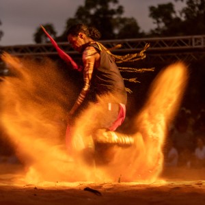 First Nations man with ochre body paint kicking up dust as he dances barefoot.