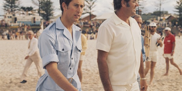 Charles, Prince of Wales wearing a blue safari suit, walking on the beach with a group of men.