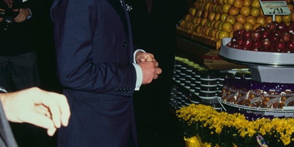 Charles, Prince of Wales standing in a supermarket in front of fruit and flowers.