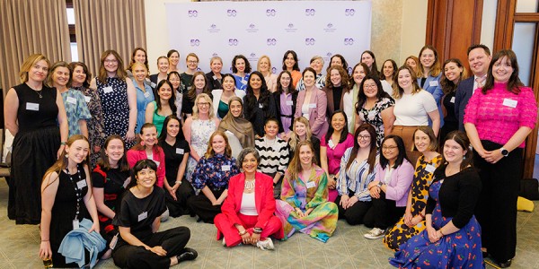 A group of 48 people pose for a group photo in front of a large purple banner printed with ‘Office for Women 50th Anniversary'.