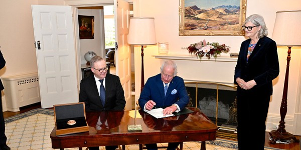 His Majesty signs a Royal Warrant. He is sitting at a desk next to Prime Minister Anthony Albanese while Her Excellency the Hon Sam Mostyn AC looks on. The new Great Seal of Australia sits in a case on the desk.