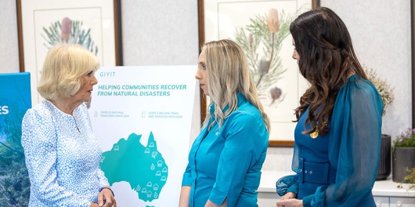 Her Majesty speaks with 2 women in front of a sign with a map of Australia and the words ‘GIVIT: helping communities recover from natural disasters’.