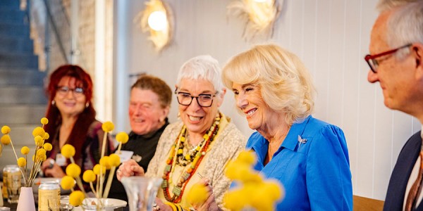 Her Majesty sits, smiling, at a table, with a plate of food in front of her. Other diners, also smiling, sit on either side.