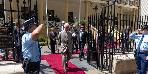 His Majesty walking out of the NSW Parliament on a red carpet being saluted by guards and followed by security.