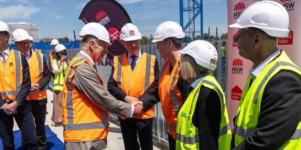 His Majesty wears a high-vis vest and a hard hat among a group of people wearing the same. He is shaking hands with one of them. 