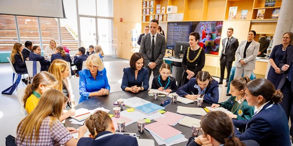 Her Majesty sits at a table with a group of children in school uniform. 