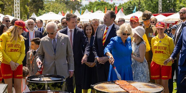 Their Majesties tend snags on barbecues while a crowd looks on. Some of the crowd are wearing Surf Life Saving Australia uniforms.