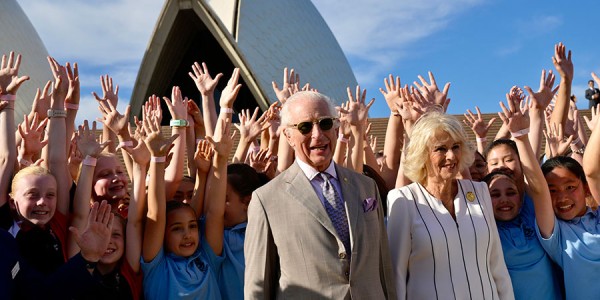 Their Majesties stand with a crowd of children in front of the Sydney Opera House. The children have their arms stretched towards the sky.