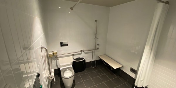 Shows inside an accessible bathroom with a toilet in the left of the room, with an accessible shower, wall mounted hand rails and with a wall mounted bench on the right.