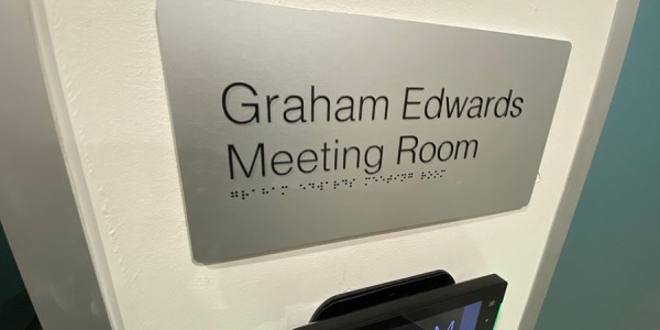 Shows a sign outside a meeting room reading “Graham Edwards Meeting Room,” with braille at the bottom of the sign.