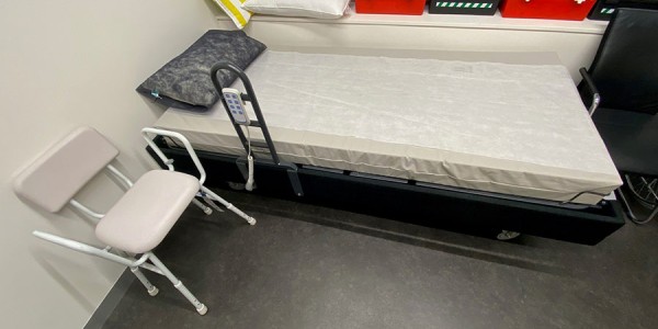 Shows a room with an electric, height adjustable bed at the far end of the room, a chair to the right of the bed, and a collection of first aid boxes.