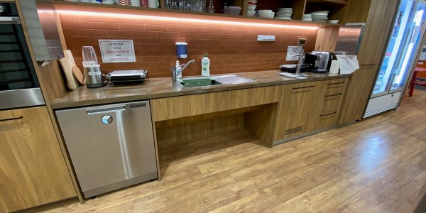 Shows a kitchen area with a refrigerator on the far right wall, with a long kitchen bench that has a wheelchair accessible sink, water station, and kitchen appliances.