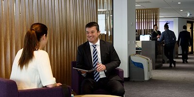 A man and a woman sit on chairs next to a small table. They both wear business clothing. In the background is a divider, a corridor and more people.