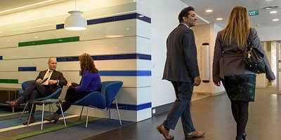 A man and a woman sit on seats in a waiting area. Another man and woman walk past them down a corridor. All are dressed in business clothing.