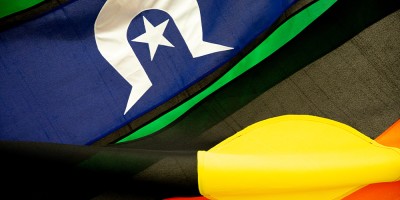 Two flags curled and together. One at top left is blue and green with a white design which includes a star. At right bottom is a flag with black, yellow and red colouring.