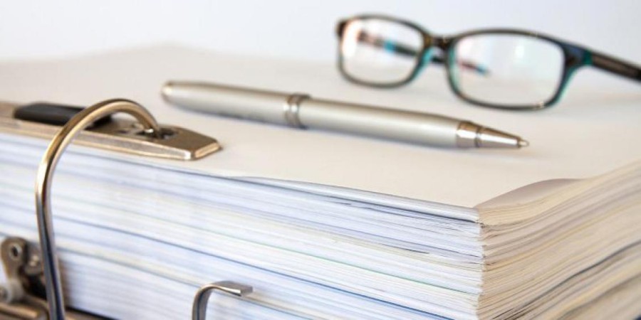 A pair of glasses and a pen sit atop an open binder showing the pages bound by a large ring.