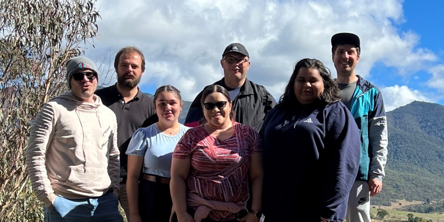 In the foreground a group of six people stand smiling and posing for a photo on top of a rocky cliff. In the background is a valley filled with mountains and trees and a slightly cloudy blue sky.