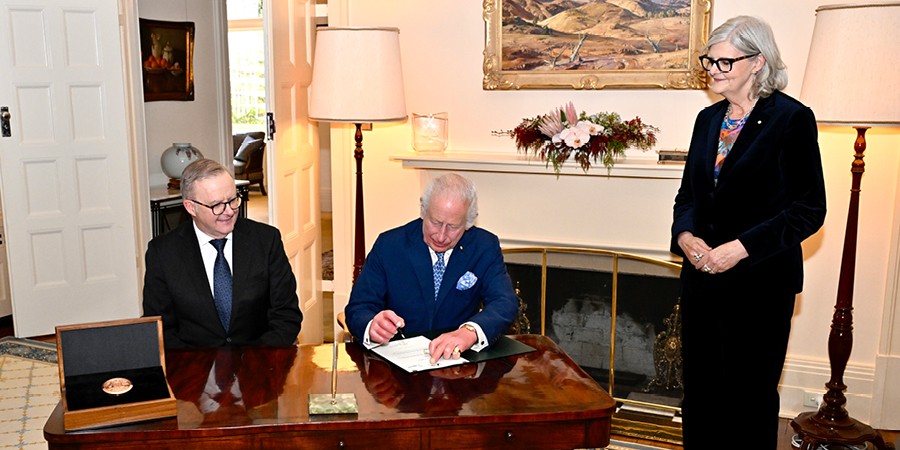 His Majesty The King Charles III signs a Royal Warrant at a desk, seated next to Prime Minister the Hon Anthony Albanese. Her Excellency the Honourable Sam Mostyn AC stands beside the desk. The new Great Seal of Australia sits in a case on the desk.